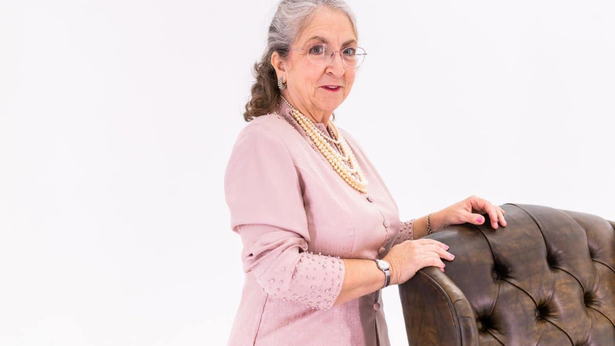 Stylish elderly woman in pink jacket standing beside an elegant chair indoors.