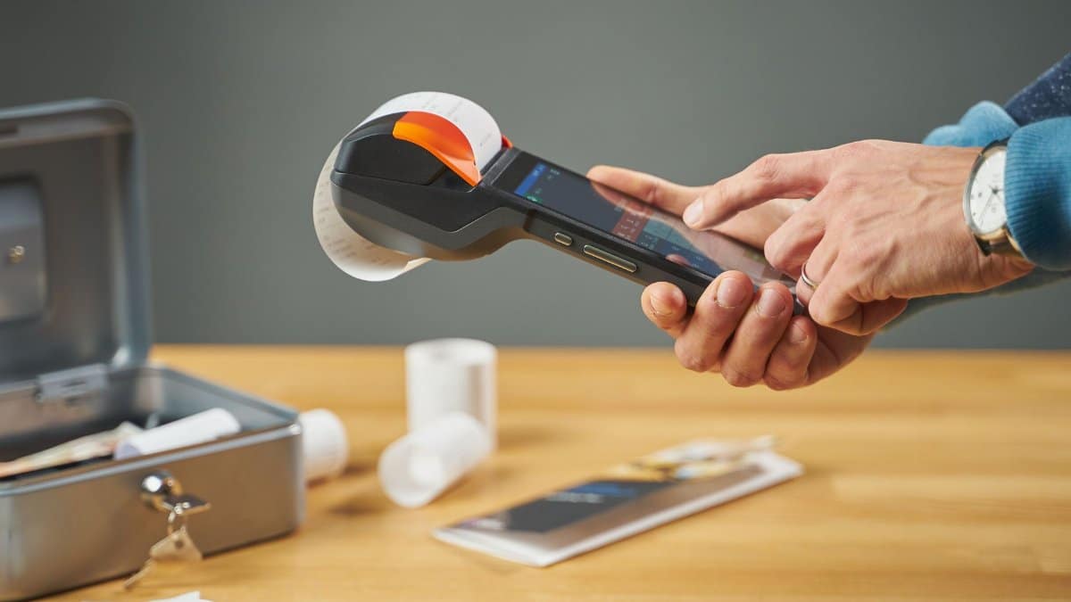Hands operating a portable receipt printer with printed receipts on a wooden table.