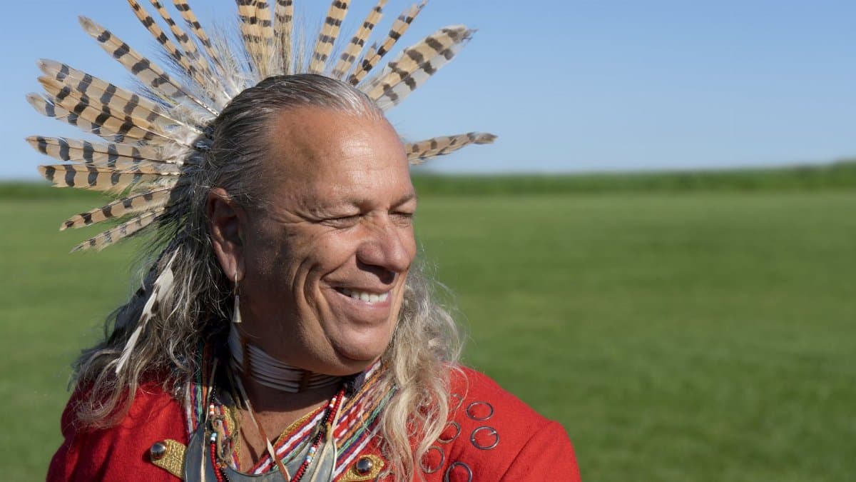 Indigenous man wearing traditional headdress and red attire, smiling outdoors in a grassy field.
