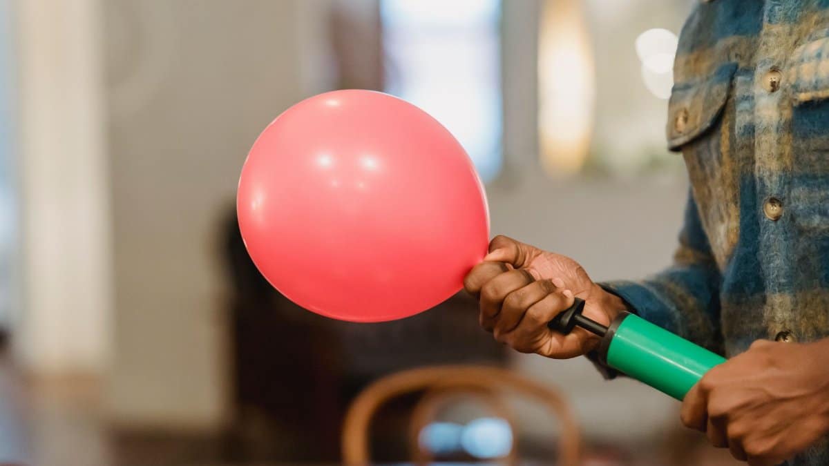 Crop faceless African American male in casual clothes holding green pump and red balloon while decorating room to festive event