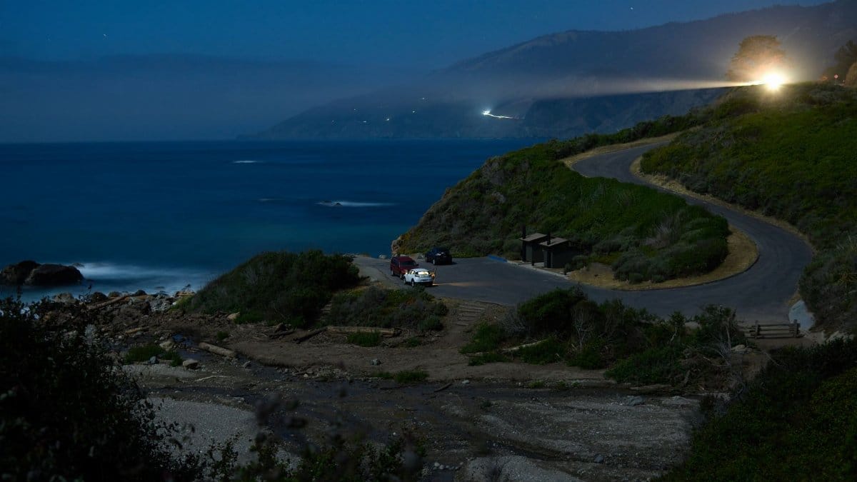 Dramatic night view of a coastal road under a star-filled sky, featuring ocean and light trails.