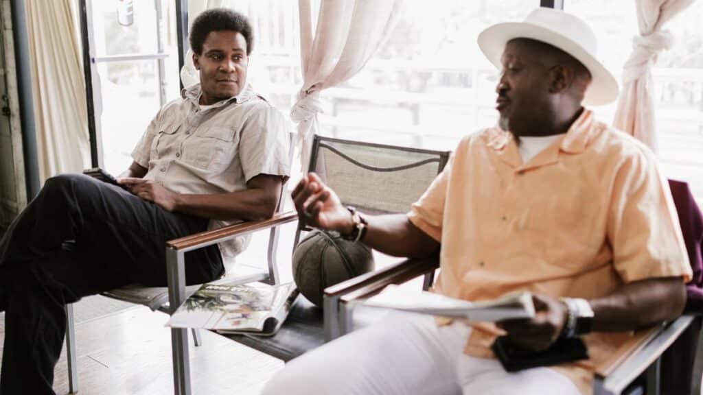 Two men engaging in a relaxed conversation inside a sunny barbershop setting.