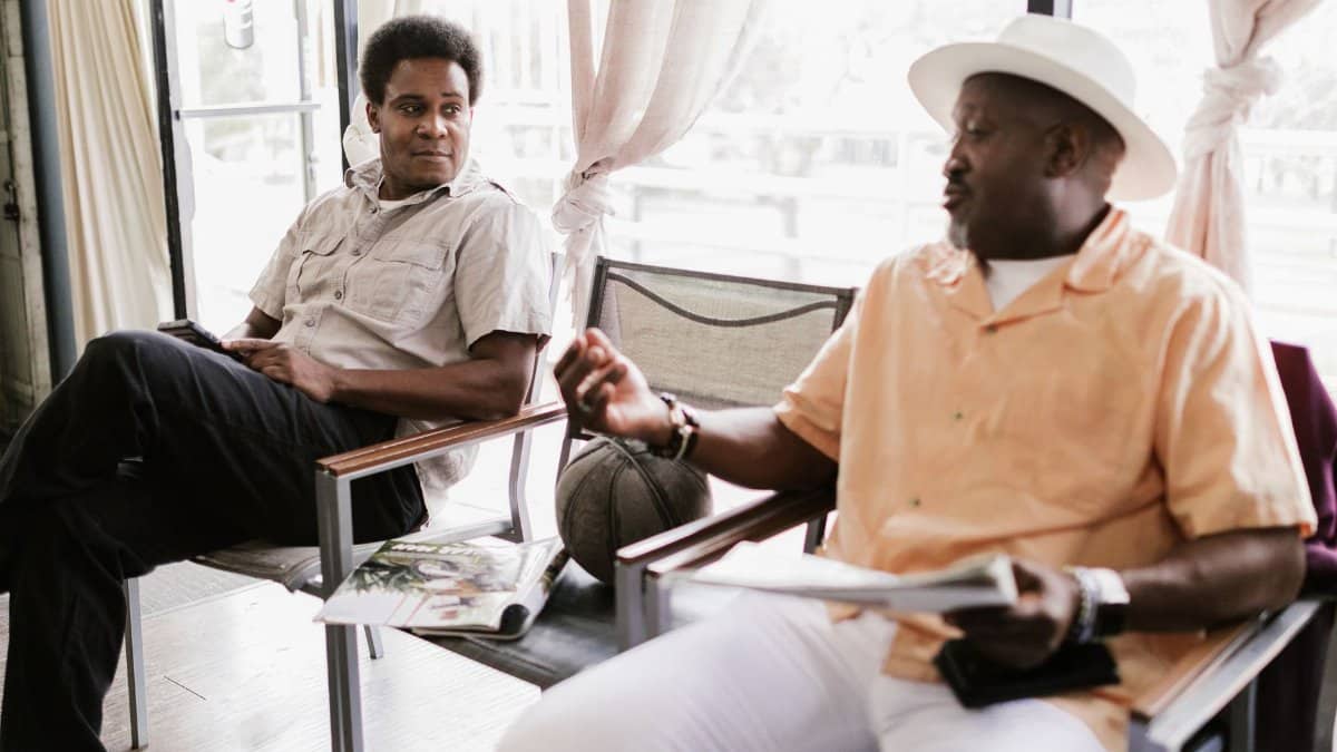 Two men engaging in a relaxed conversation inside a sunny barbershop setting.