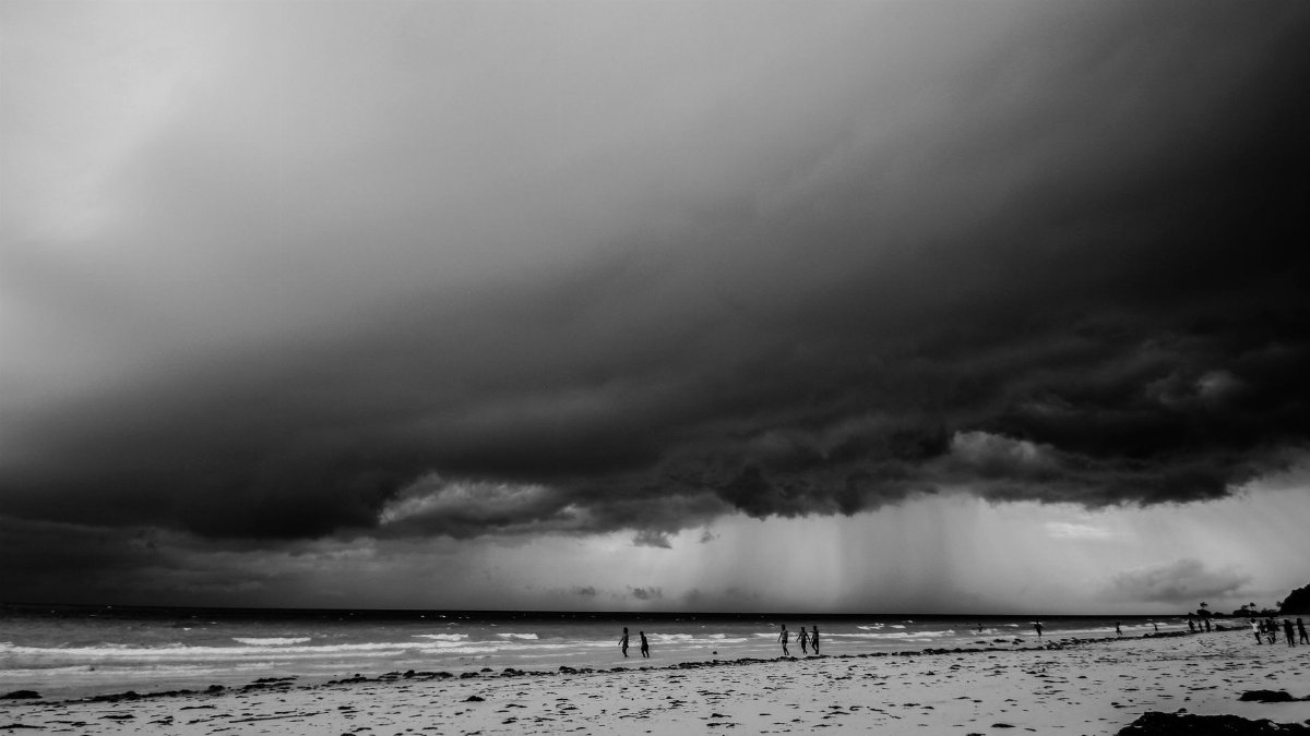 Monochrome view of an impending storm over a beach with people walking along the shore.