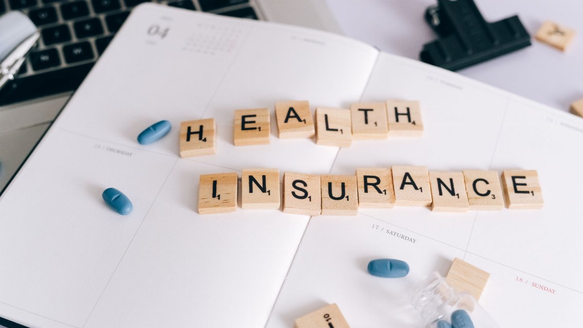 Scrabble tiles spelling 'Health Insurance' on planner with pills and laptop, symbolizing healthcare planning.