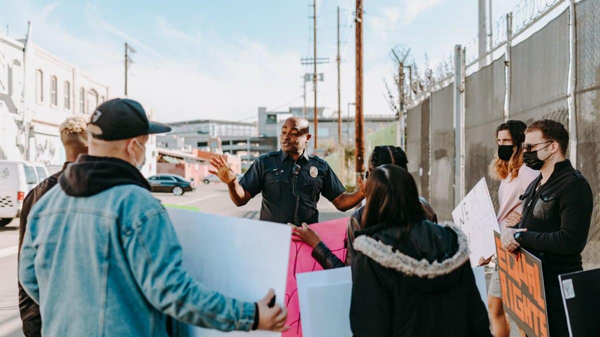 A police officer addresses a group of peaceful protesters holding signs on a city street under a clear sky.