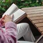 Elderly woman in pink shirt reading book outdoors at wooden table, enjoying leisure time.
