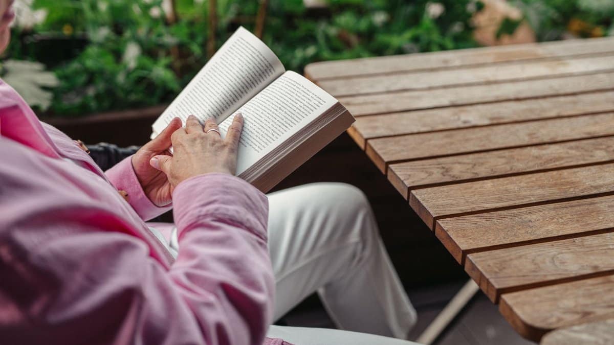 Elderly woman in pink shirt reading book outdoors at wooden table, enjoying leisure time.