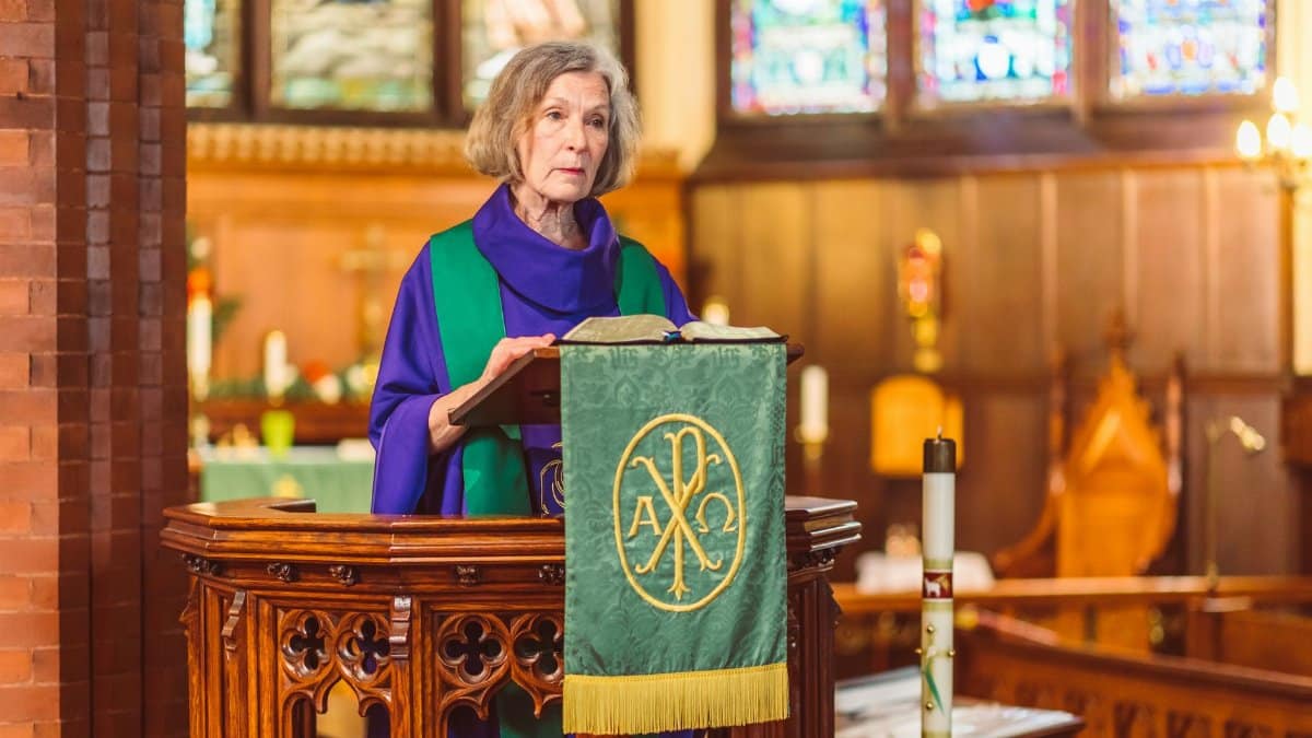 A senior female priest speaks at a pulpit in a beautifully lit church interior.