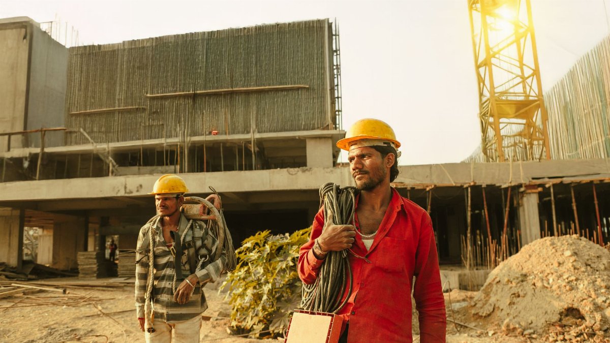 Two construction workers wearing helmets at a site in India with a crane and building.