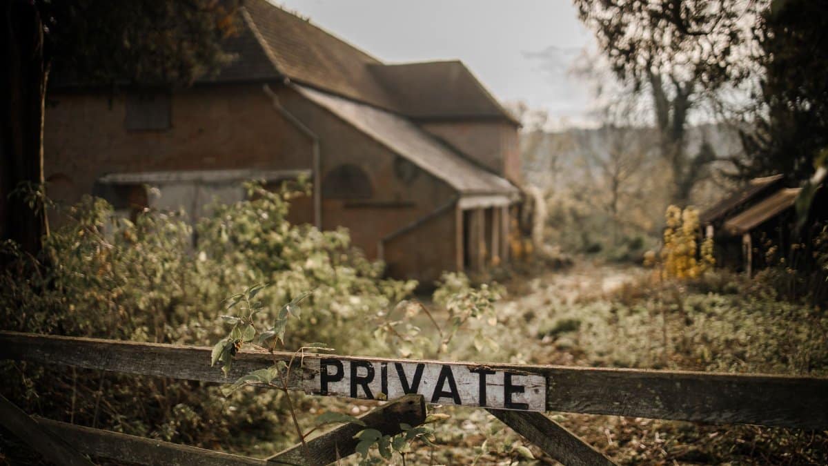 Rustic wooden gate with a 'PRIVATE' sign leading to an old countryside house, surrounded by greenery.