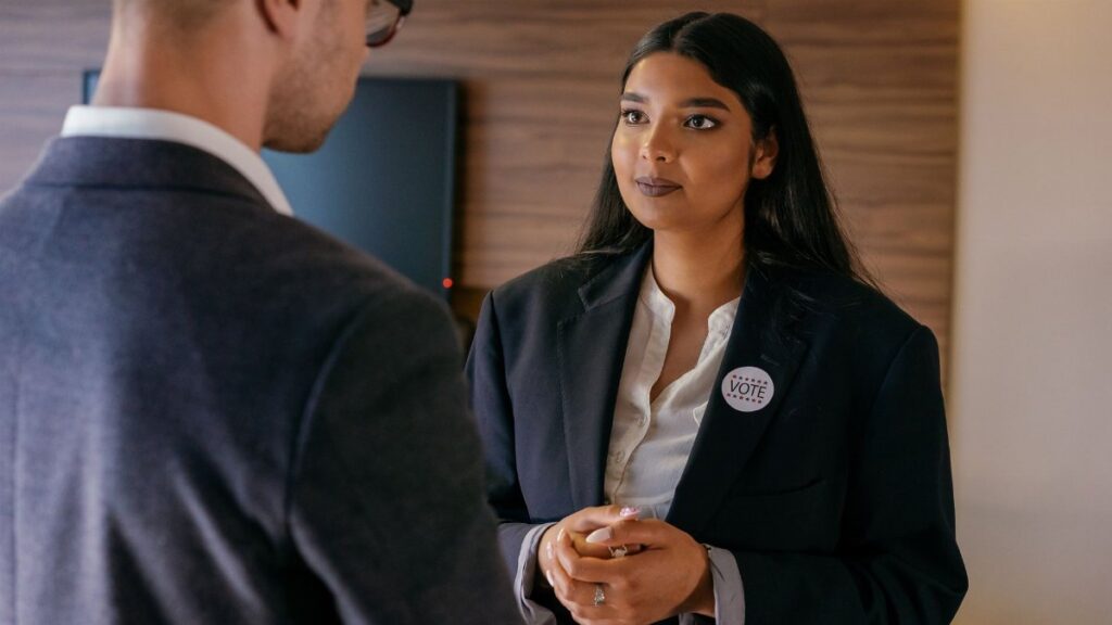 A female candidate in a political discussion, wearing a vote badge during an election campaign indoors.