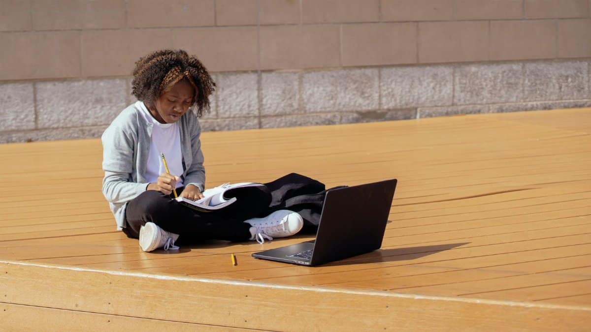 Full body of focused African American girl sitting with laptop and copybook and doing homework