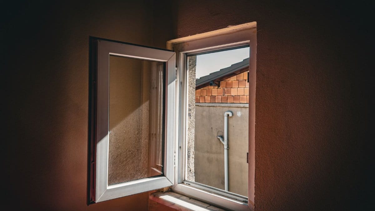 Open window view of a brick wall and pipe, sunlight casting shadows indoors.