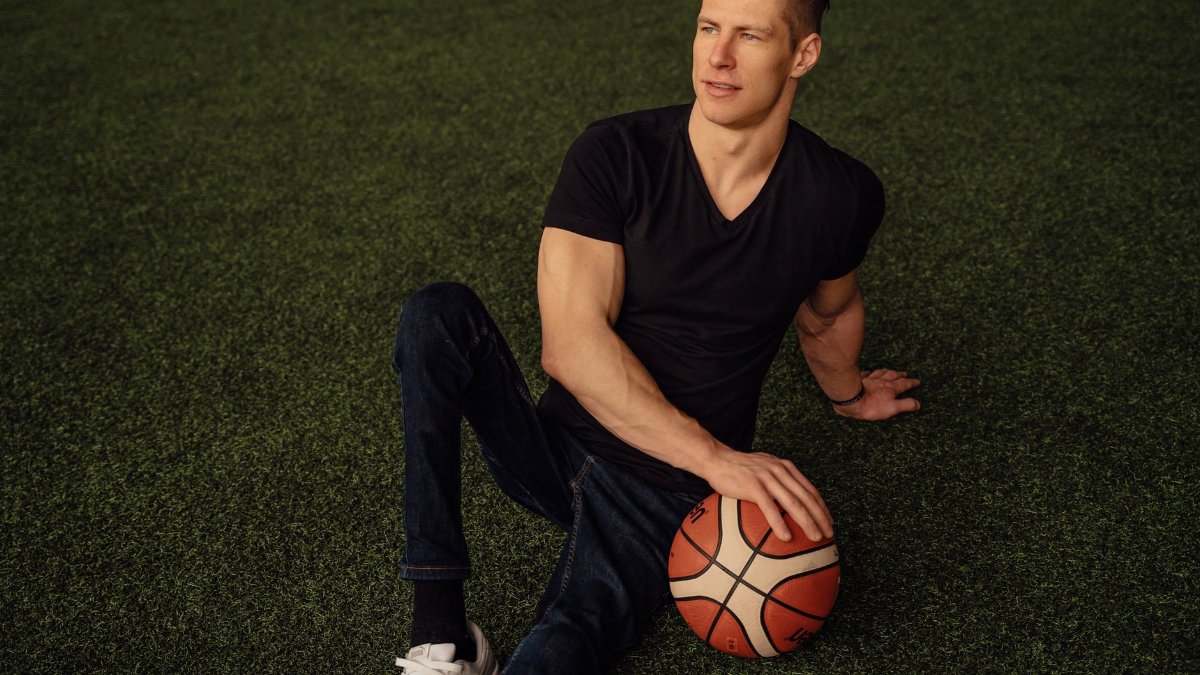 A man sits on artificial grass holding a basketball, dressed in casual attire and looking thoughtfully away.