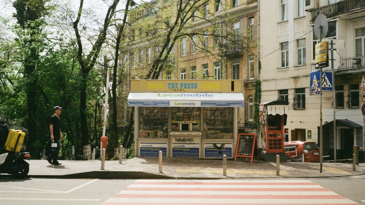 Urban street scene in Kyiv, featuring a magazine stand, residential buildings, and a passing pedestrian.