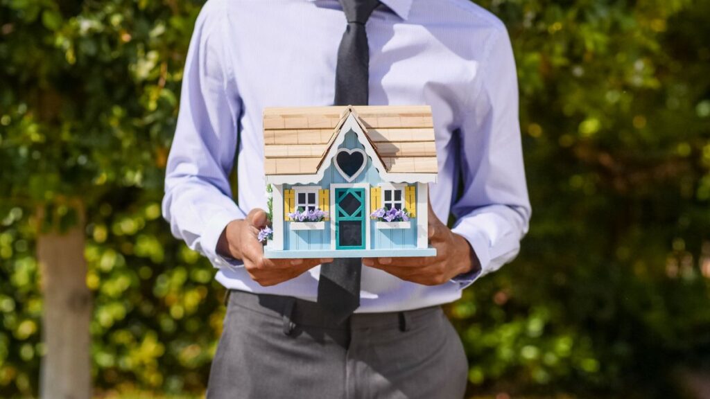 A business professional holds a decorative miniature house, symbolizing real estate investment.