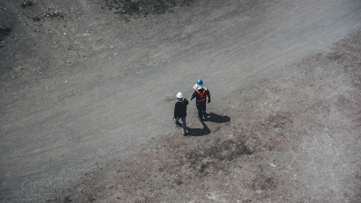 Two construction workers walking on a gravel road with safety gear and helmets, seen from above.