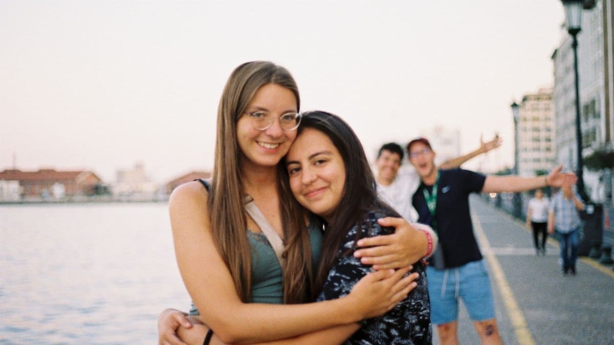 Two young women embrace happily by a waterfront with friends in the background.