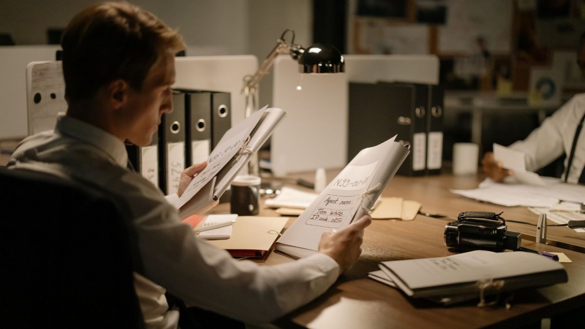 Two detectives reviewing documents in a dimly lit office setting, focused and professional.