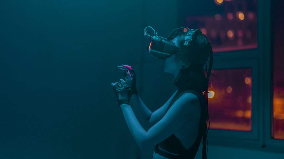 A woman wearing a VR headset and gloves immersed in a virtual reality experience inside a dimly lit room.