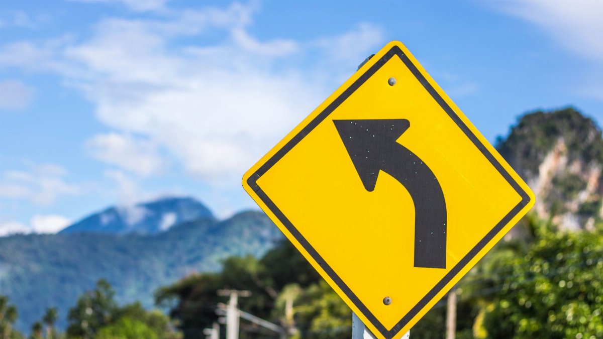 Bright yellow left turn traffic sign with mountain landscape in the background outdoors.