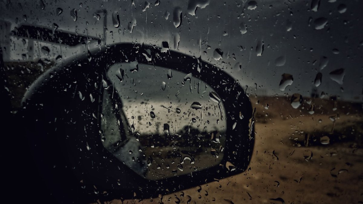 Close-up of a car side mirror with raindrops, capturing a wet and stormy atmosphere.