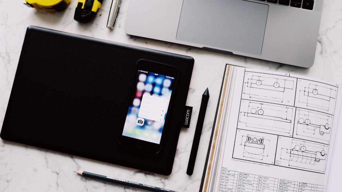 From above view of modern workplace with laptop and tablet and smartphone placed near opened notebook with drawings and pen with pencil on white marble table
