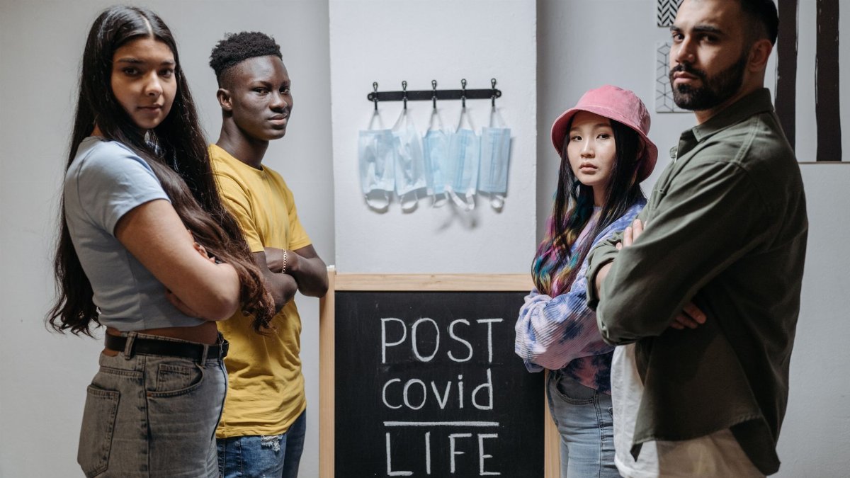 Diverse individuals standing in front of a chalkboard reading 'Post COVID Life' with hanging face masks.