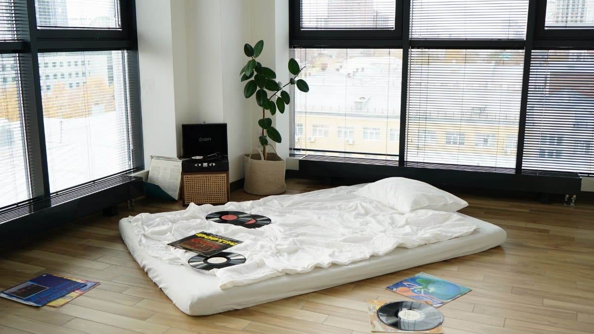 A minimalist apartment interior with a mattress on the floor surrounded by vinyl records and a record player.