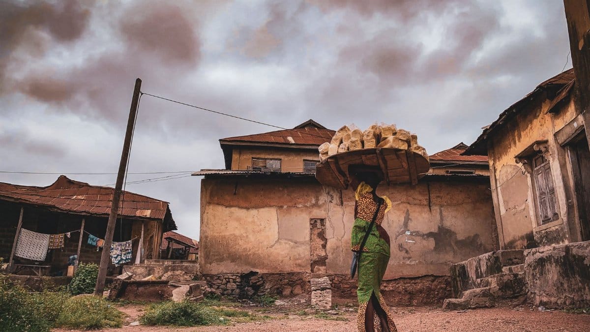 A rural village scene featuring a woman carrying goods on her head in traditional attire.