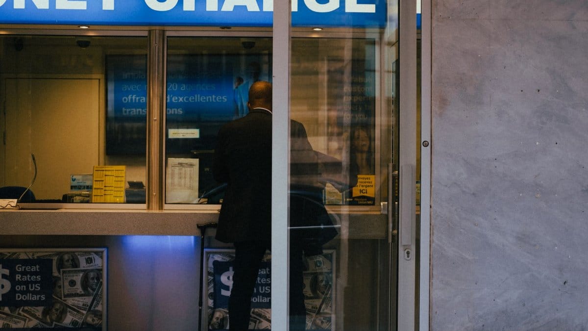 Man at a currency exchange office window, showing currency rates inside a bustling city.