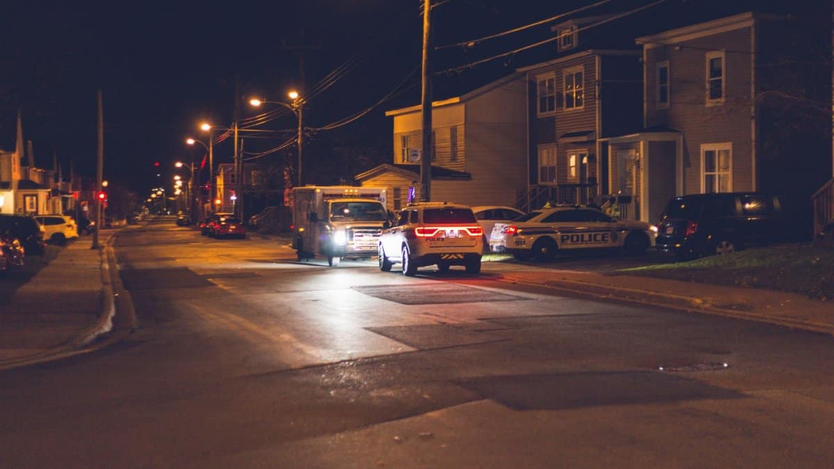 Cars with glowing lights parked on asphalt road near police car placed near residential building in small town in evening