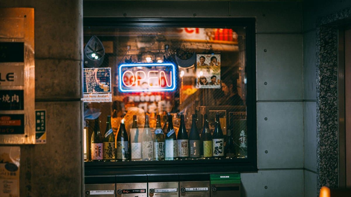 A cozy Japanese shop window displaying sake bottles and a vibrant neon open sign, evoking night ambiance.