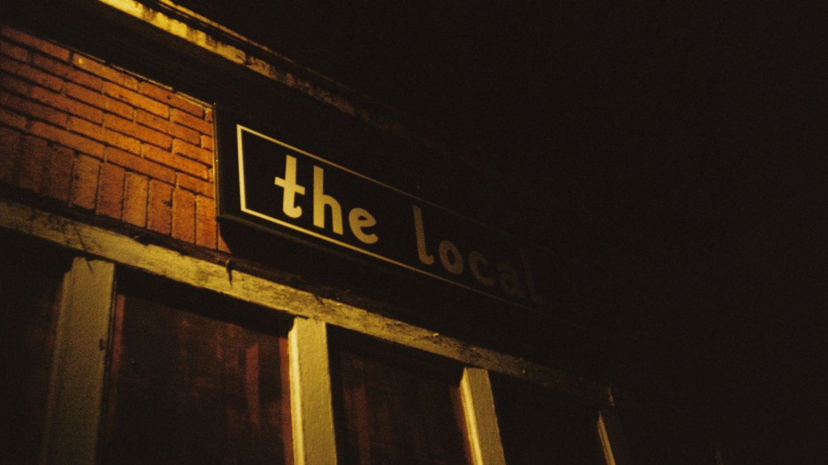 A dark and moody photo of the facade of 'The Local' bar at night, with warm lighting.