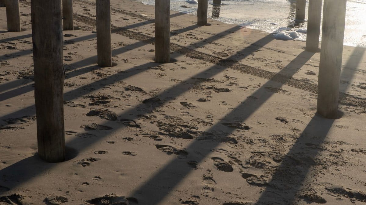 Long shadows cast by pier posts on a sandy sea beach during sunset.