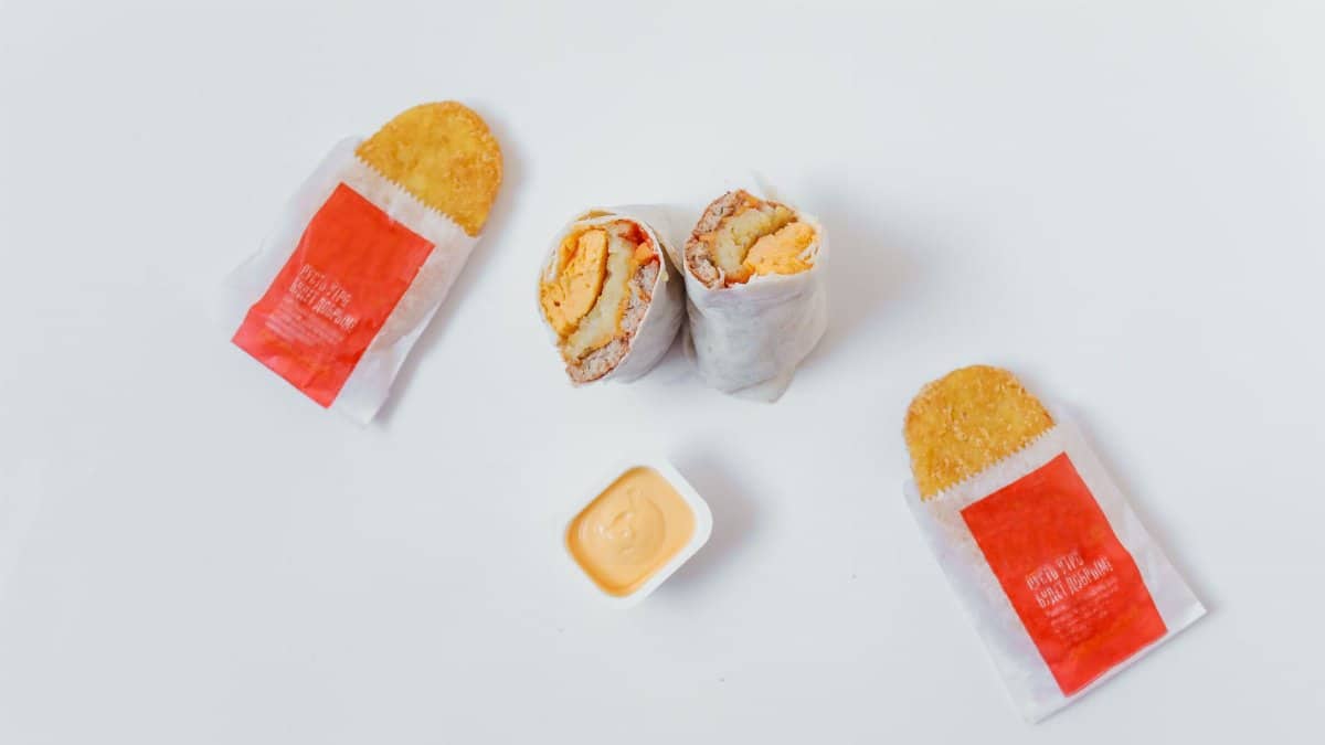 Flat lay of breakfast burrito and hash browns with sauce on white background.
