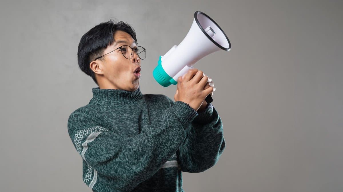 Portrait of an Asian man wearing glasses and a sweater, dynamically using a megaphone indoors.