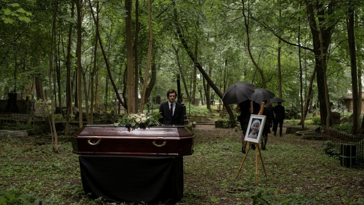 A funeral ceremony in a wooded cemetery with a wooden coffin and mourners.