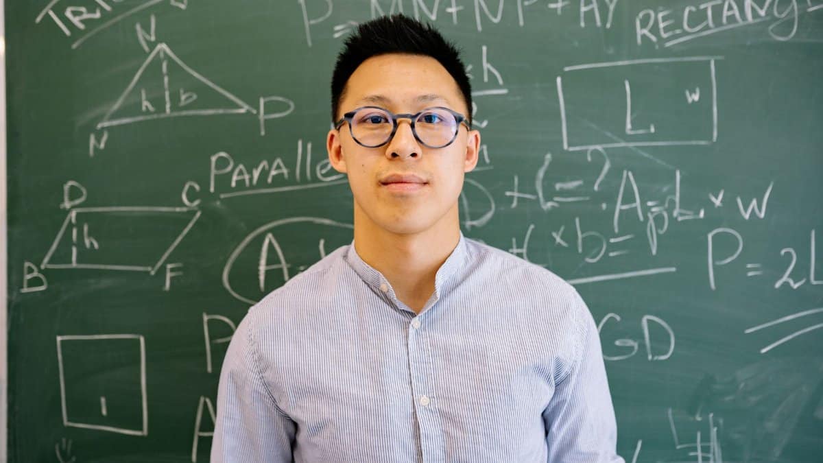 Educator standing before chalkboard filled with geometry and algebra equations, wearing glasses and long-sleeved shirt.