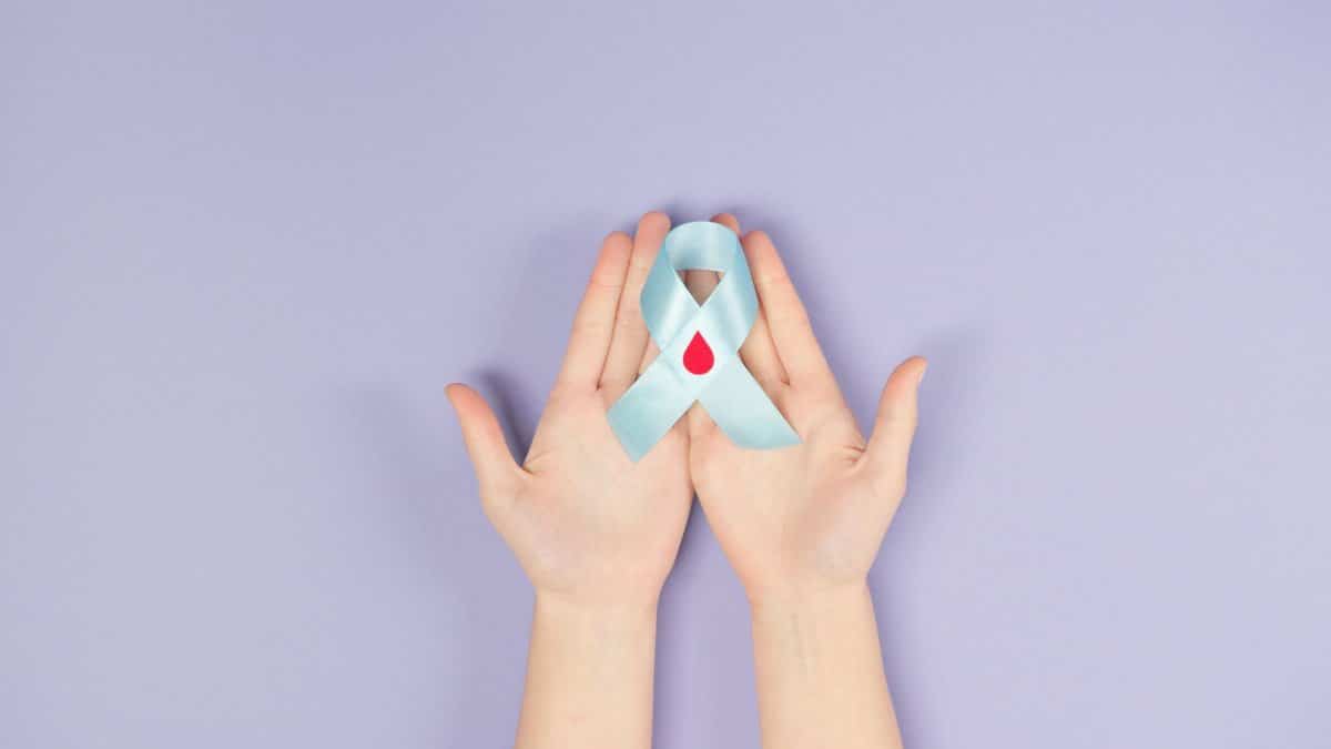 Hands holding a blue ribbon with a red blood drop symbolizing diabetes awareness against a purple background.