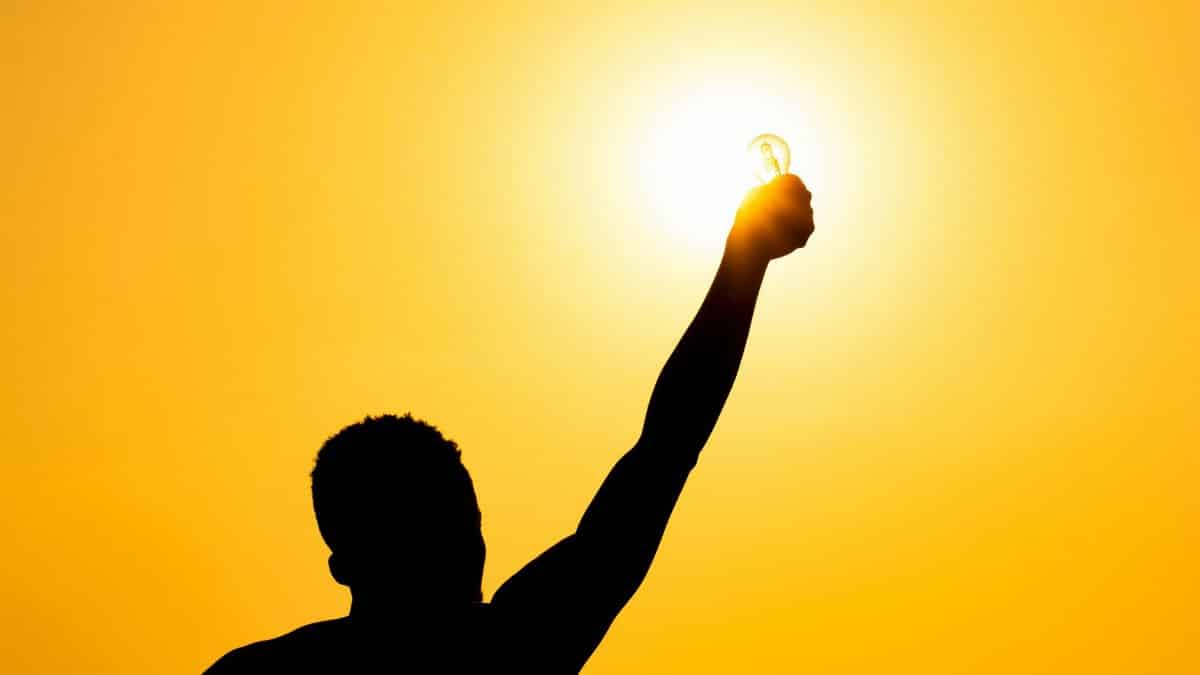 From below of silhouette of faceless male raising hand with light bulb against bright sundown sky