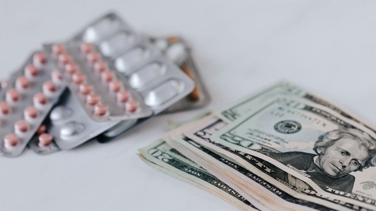 Heap of similar American dollars in cash placed near blisters of expensive drugs on white table