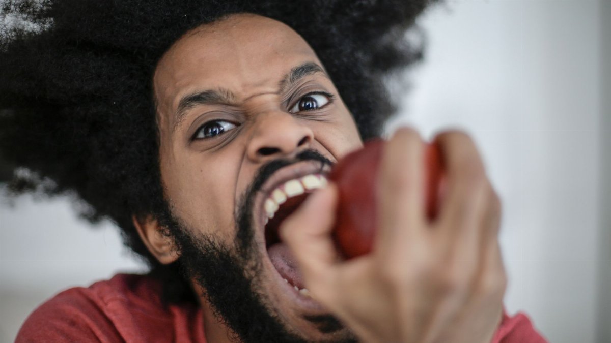 Energetic portrait of a man with afro hair biting into a red apple with an intense expression.