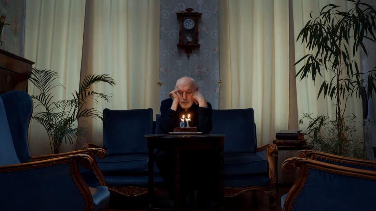 Senior man alone with a birthday cake in a vintage living room setting.