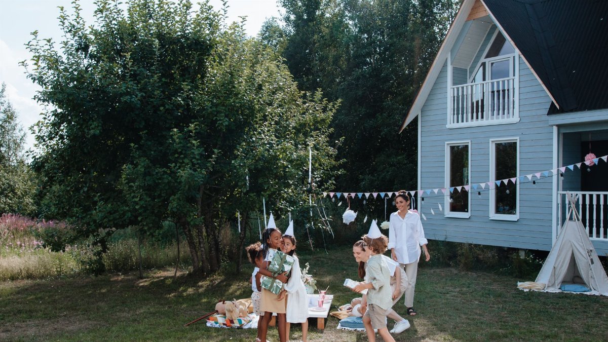 Children playing at a summer outdoor party beside a cozy house with nature backdrop.