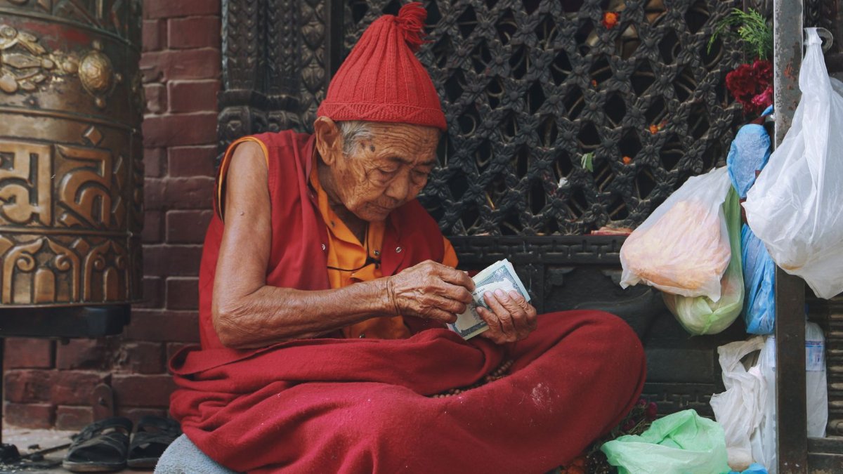 An elderly monk sits cross-legged on a street, counting money amidst bags and traditional Tibetan attire.