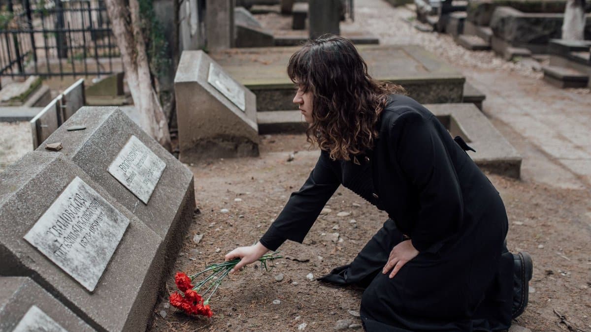 A woman in a black dress kneels by a tombstone with red flowers, expressing loss and remembrance.