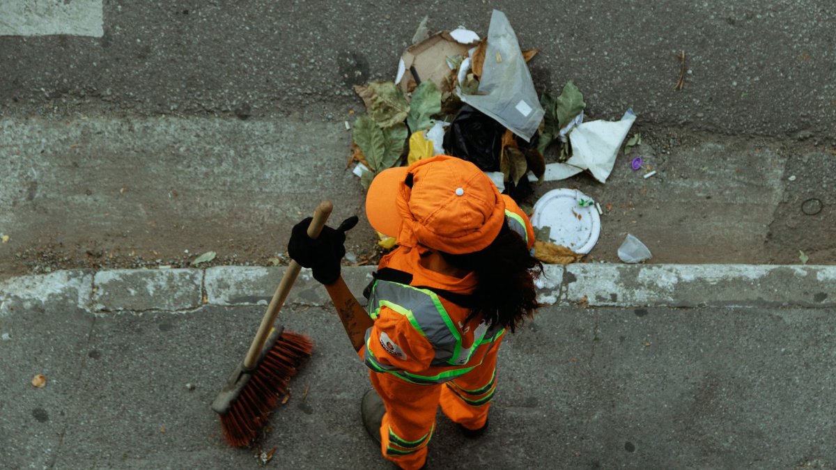 Top view of a street cleaner wearing orange uniform sweeping debris on a city sidewalk.