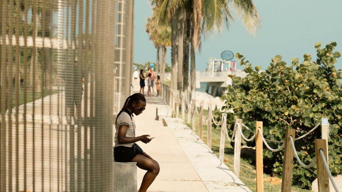 A woman sits and relaxes along a sunny Miami waterfront promenade, palm trees lining the path.